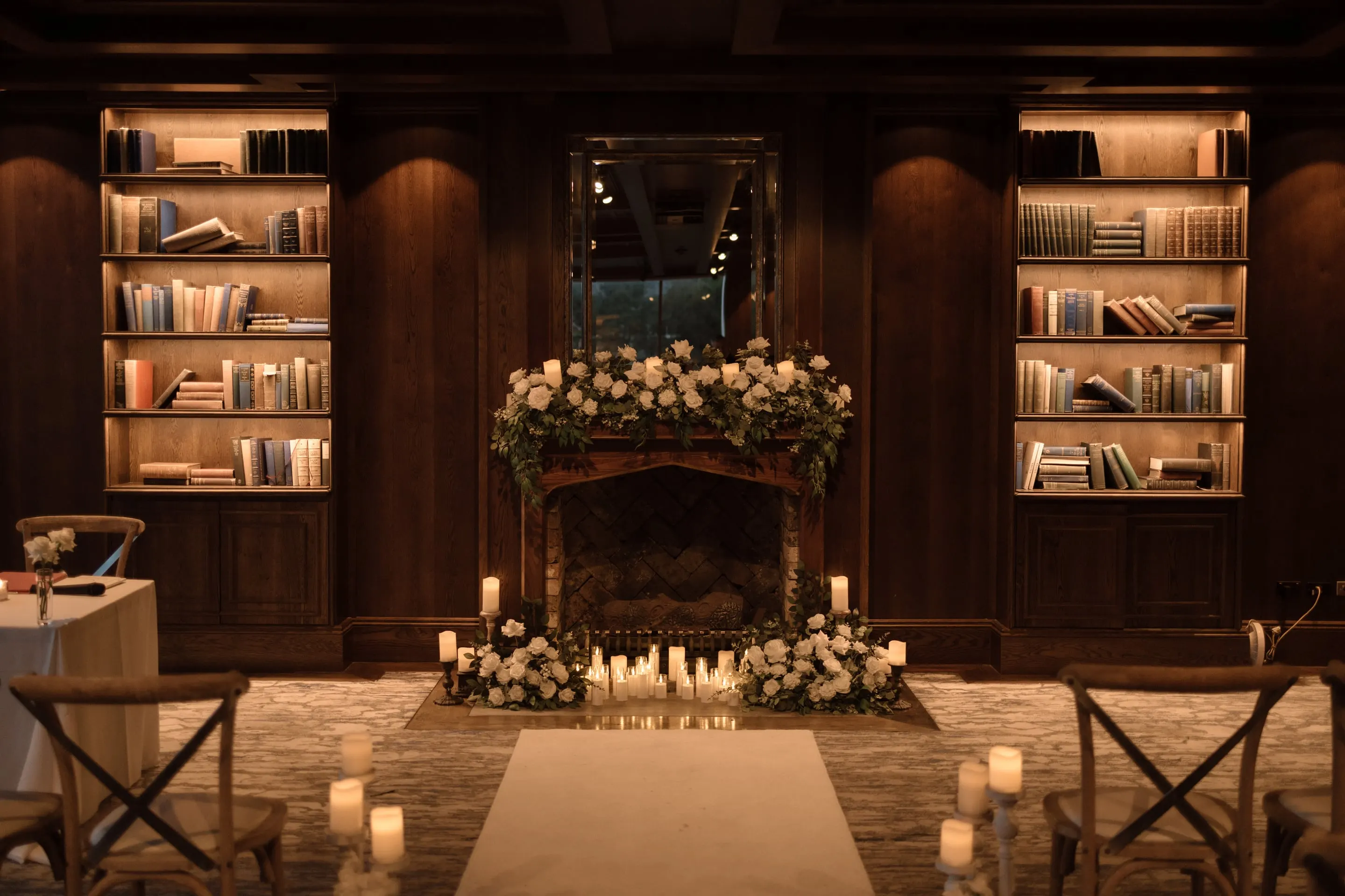 Candlelit ceremony aisle between timber chairs, leading to a fireplace dressed in white roses beneath lit bookshelves